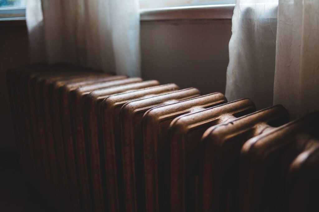Aged heating battery made of cast iron near windowsill with curtains in house