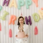 Woman celebrating birthday with colorful balloon decorations and a cake indoors.