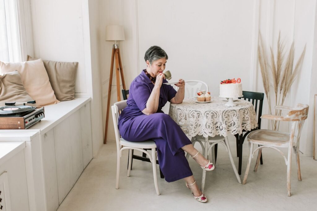 A senior woman gracefully celebrates her birthday in a stylish indoor setting with cake and tea.