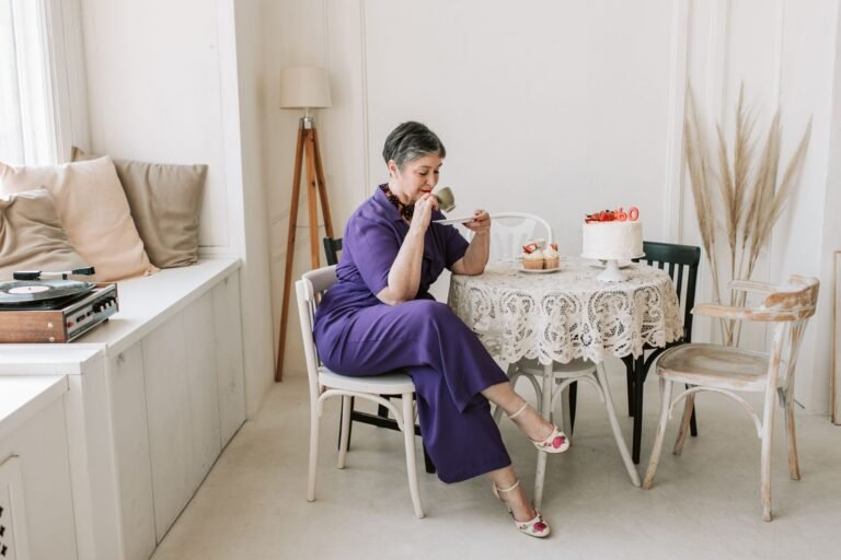 A senior woman gracefully celebrates her birthday in a stylish indoor setting with cake and tea.