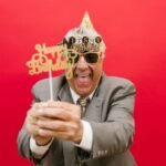 Elderly man in a suit celebrating his birthday with festive props and a red backdrop.