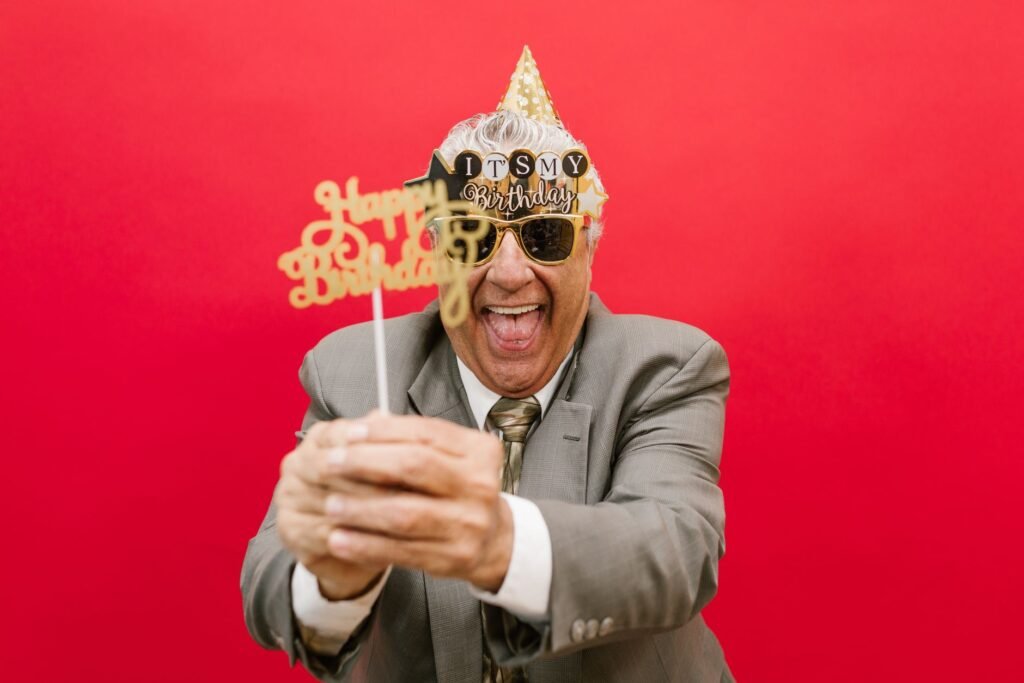 Elderly man in a suit celebrating his birthday with festive props and a red backdrop.