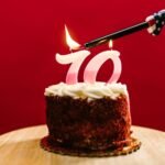 A close-up of a birthday cake with '70' candles being lit against a red background.