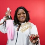 A smiling woman celebrates with a wine bottle and birthday sign against a vibrant red background.