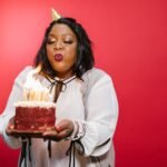A black adult woman joyfully blowing out birthday candles on a red velvet cake against a red backdrop.