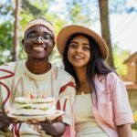 Two adults celebrating a birthday outdoors with a cake adorned with candles, smiling and happy.