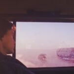 A man wearing a beanie hat sits in a vehicle, gazing through a frosty window at a winter landscape.