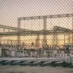View of an industrial power plant through a chain-link fence at sunrise, symbolizing energy and industry.