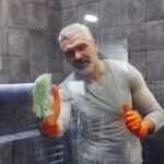 Grey haired male with beard in orange rubber gloves attentively cleaning shower glass in bathroom